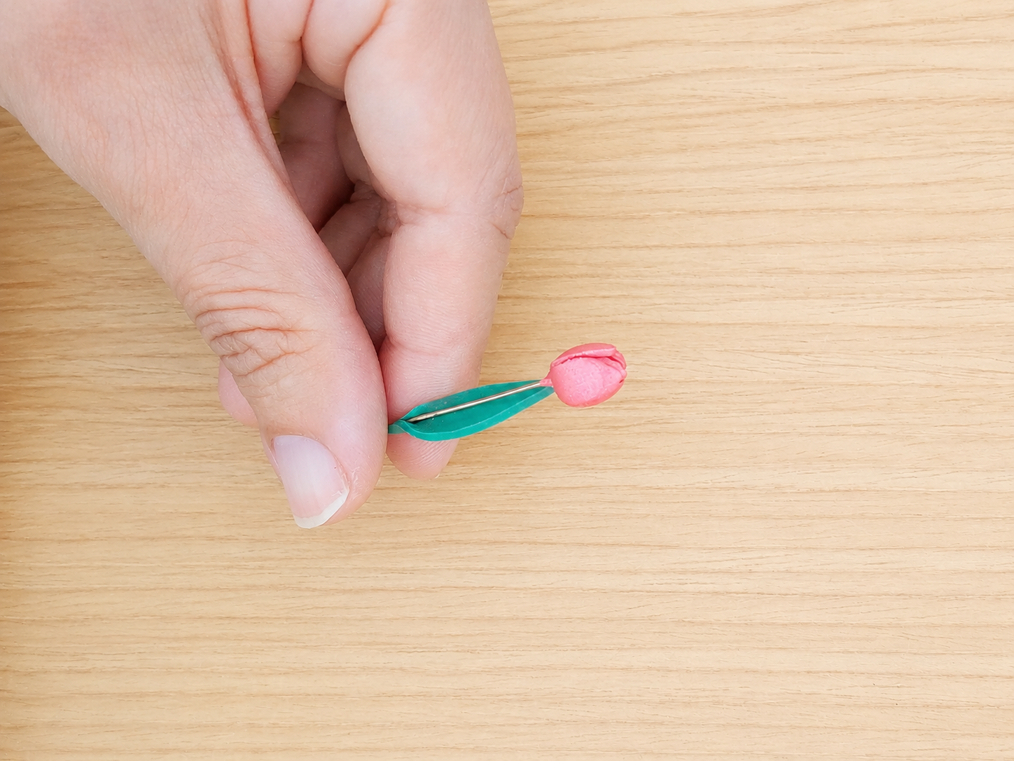 A hand holding a small pink flower-shaped accessory with a green leaf-like base over a light wood grain surface.