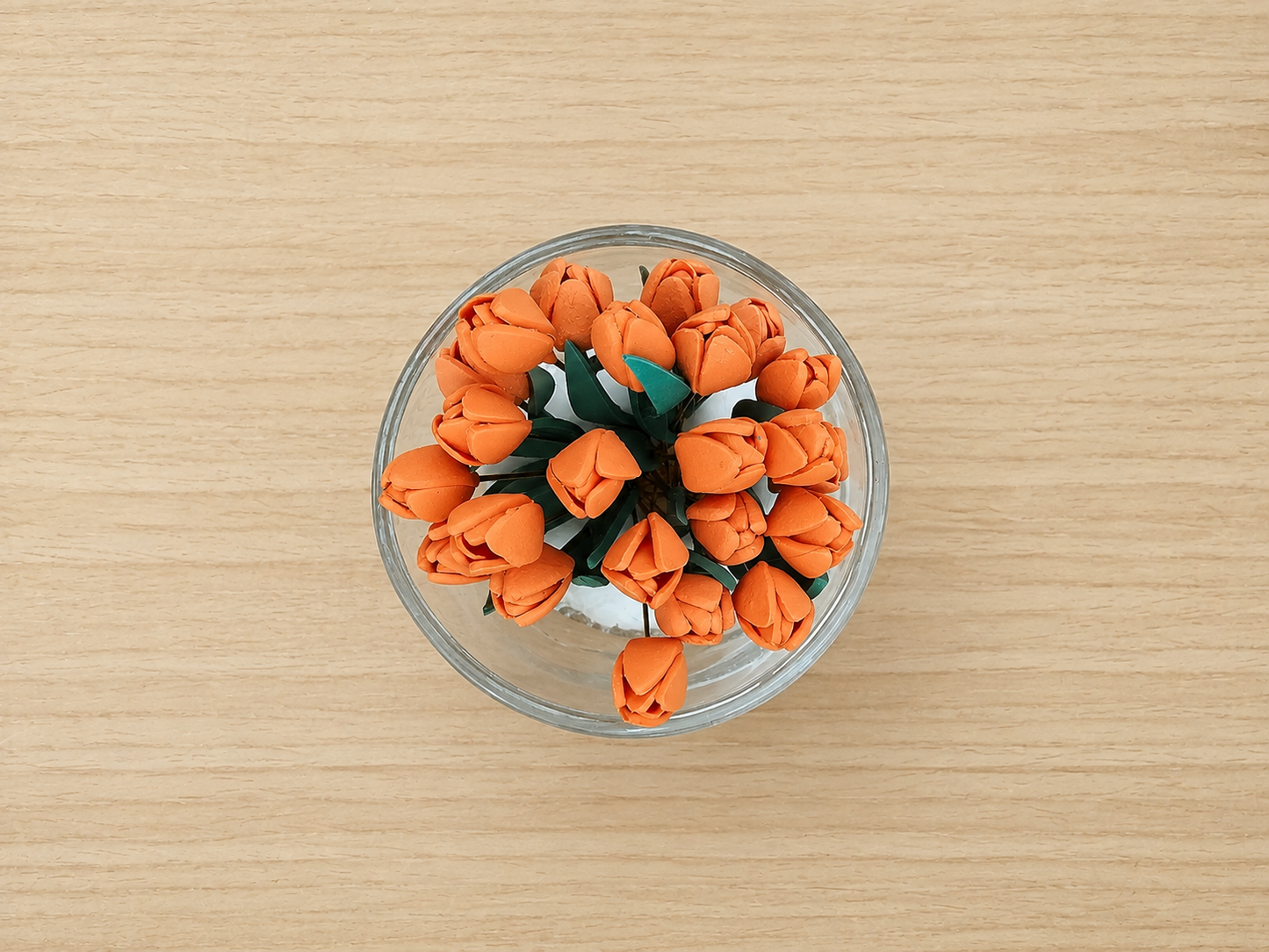 A top-down view of a small clear glass bowl filled with orange artificial tulip flowers and green leaves, placed on a light wood surface.