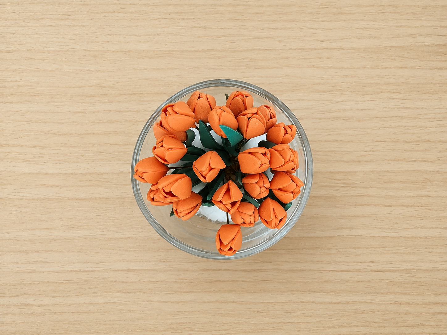 A top-down view of a small clear glass bowl filled with orange artificial tulip flowers and green leaves, placed on a light wood surface.