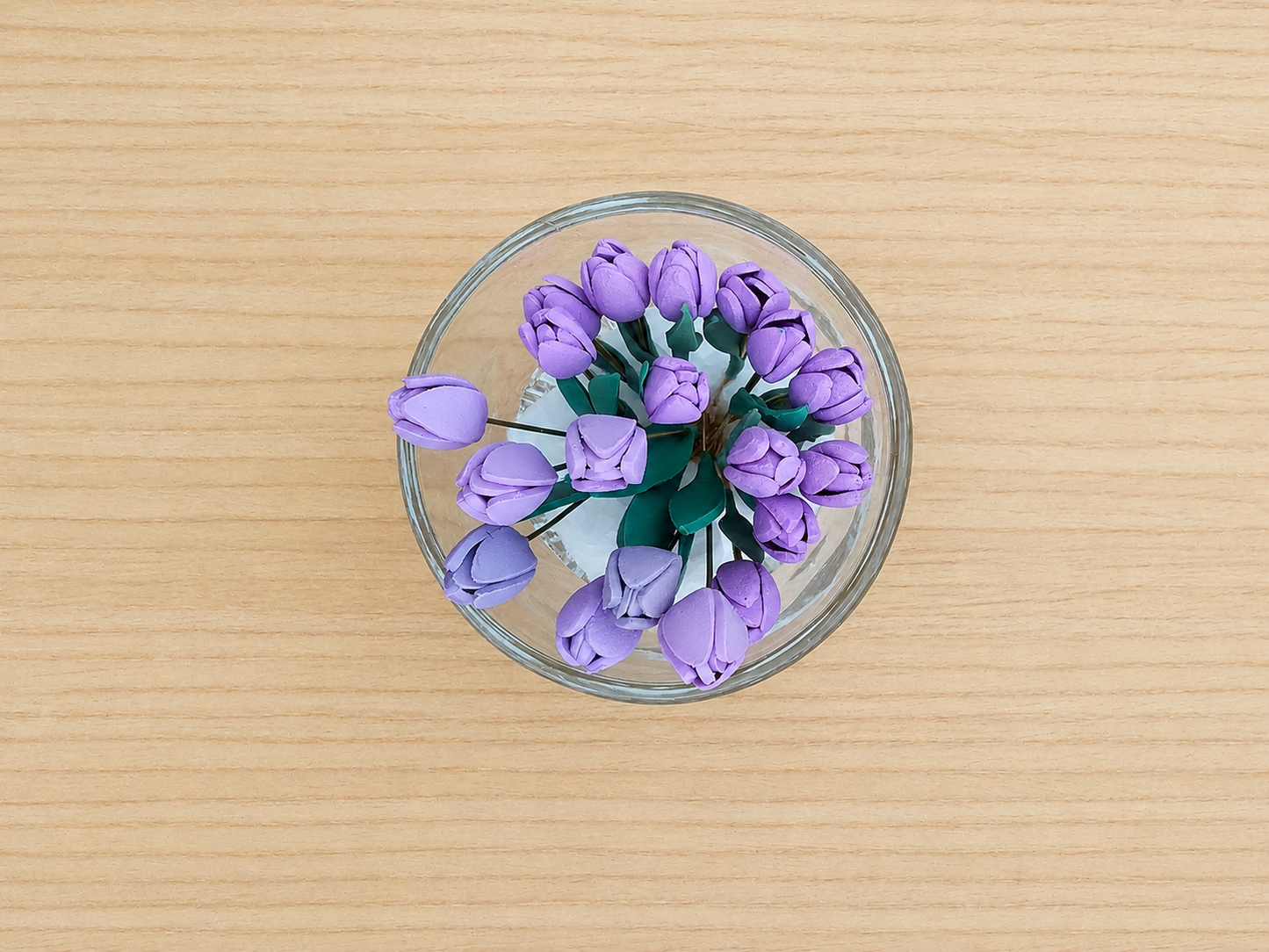 A top-down view of a clear glass bowl filled with small purple tulip-like flowers on a light wood grain surface.