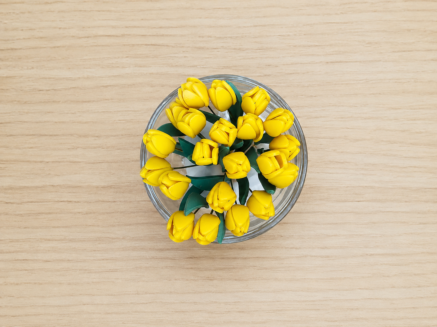 A top-down view of a clear glass bowl filled with yellow tulip-shaped ornaments or flowers with green leaves, set on a light wood grain surface.