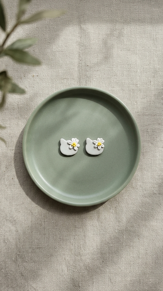 Cat-shaped earrings with flower details on a green dish against a textured beige background