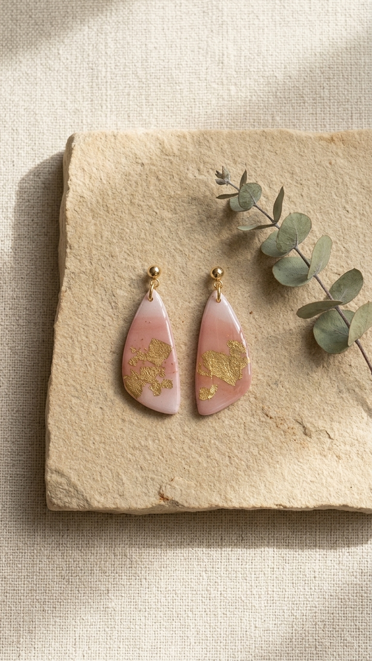 Pink and gold earrings on a stone surface with eucalyptus leaves.
