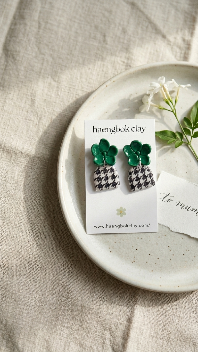 Green and black patterned earrings on a card with 'haengbok clay' branding, placed on a beige fabric background.