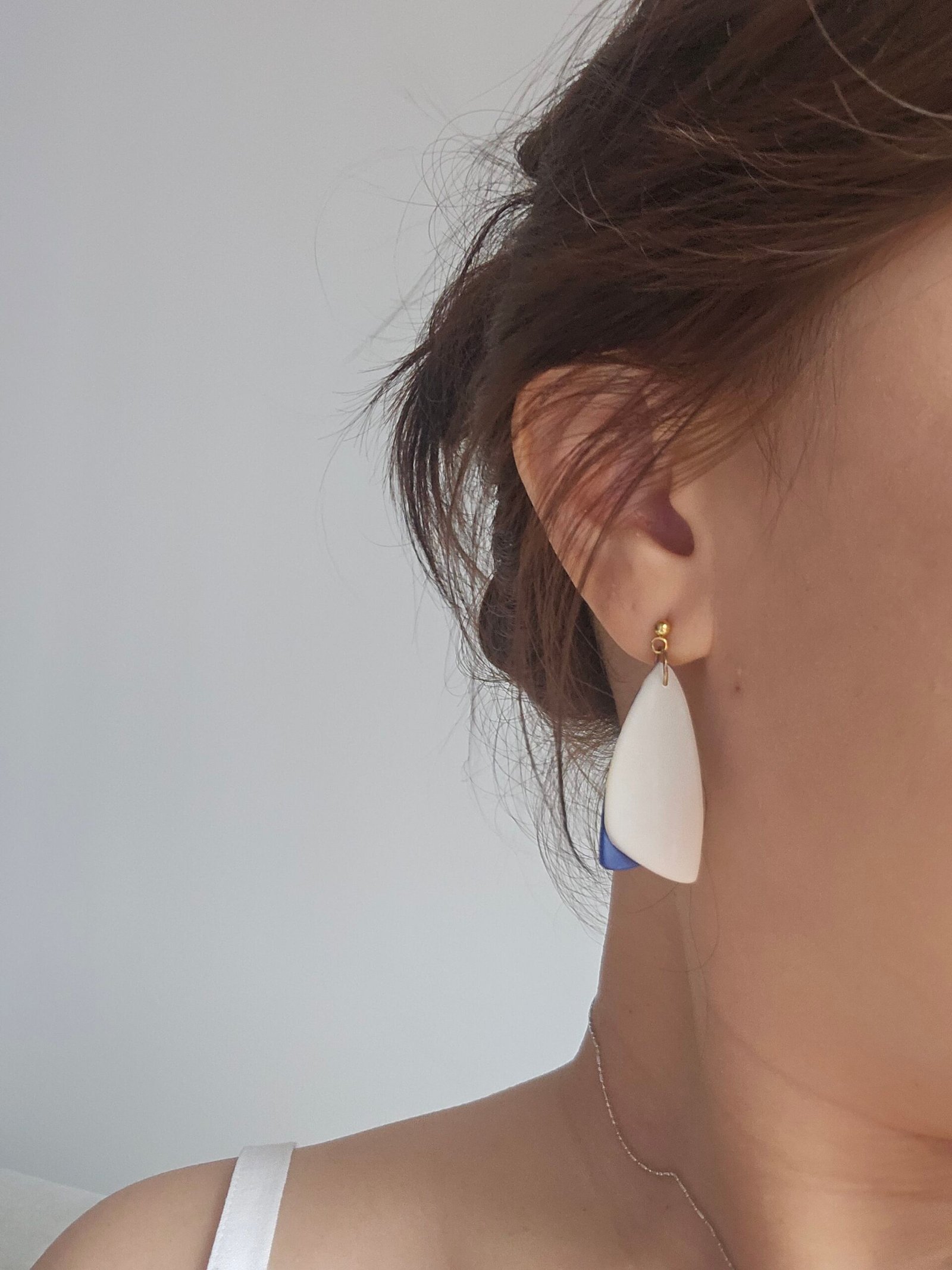 Close-up of a person wearing minimalist handmade polymer clay earrings with a white petal-shaped drop and a subtle blue accent, finished with a small gold-tone stud, against a neutral background.