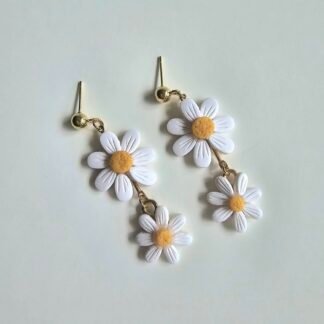 Pair of white double-daisy polymer clay earrings with yellow centres on gold-plated posts, photographed on a soft neutral background.