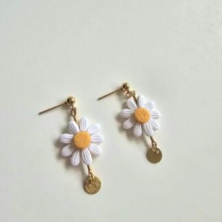 Handcrafted white daisy drop earrings with textured petals, yellow centres, and a small gold charm, set on gold-plated posts against a soft neutral background.