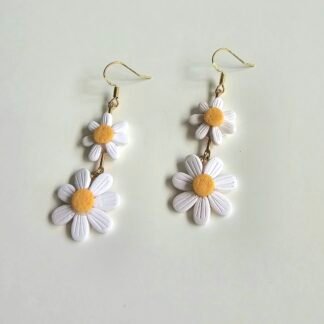 Handmade white double daisy polymer clay drop earrings with yellow centres and gold-plated hooks on a neutral background.