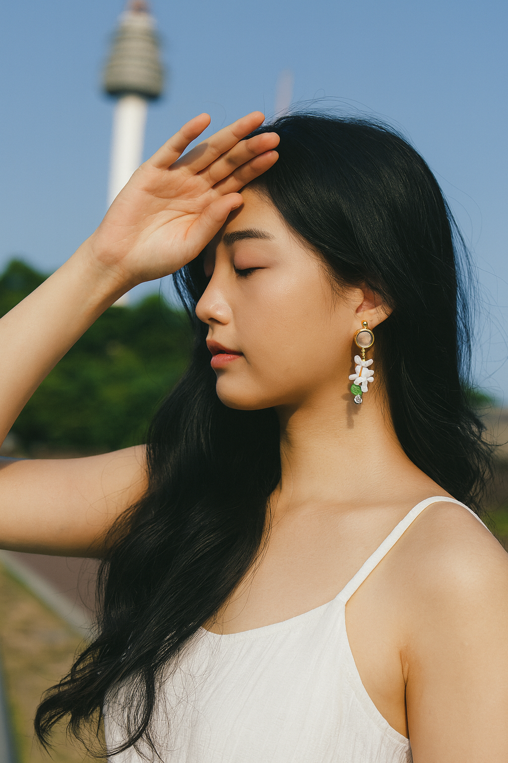Woman wearing delicate floral drop earrings outdoors, gently shading her eyes in soft sunlight, showcasing handcrafted Haeng Bok Clay jewellery in a calm, natural setting.