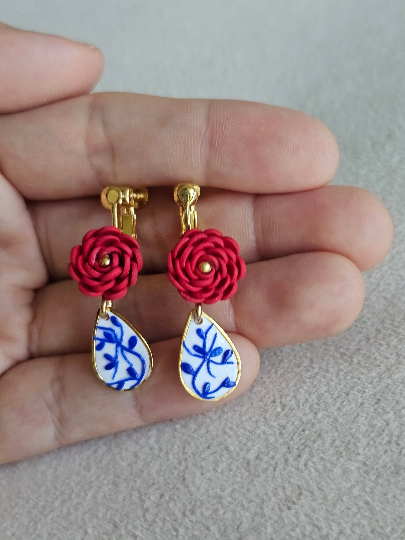 Handcrafted clip-on earrings featuring deep red polymer clay rose blooms paired with blue-and-white porcelain-style teardrop charms, set in gold-toned hardware and held against a neutral fabric background.