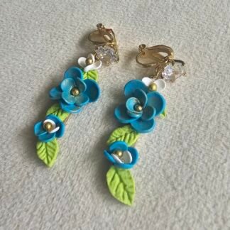 Handcrafted blue floral polymer clay clip-on earrings with green leaves and gold accents, featuring 14K gold-plated clip-on hardware on a neutral fabric background.
