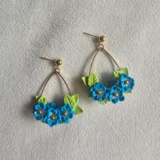Handcrafted polymer clay blue floral earrings with green leaf accents suspended on a gold arc frame against a neutral fabric background