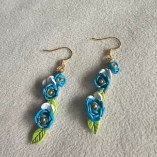 Handcrafted blue floral polymer clay long drop earrings with green leaves and white accents, finished with gold-plated 925 silver hooks on a neutral fabric background.