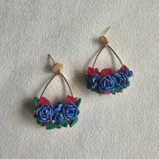 Handmade floral hoop earrings featuring blue rose clusters with red and green leaves on gold-plated posts, photographed on a neutral textured background.