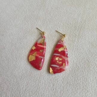 Handmade red polymer clay drop earrings with gold foil accents on gold-plated posts, photographed resting on a hand against a neutral fabric background.