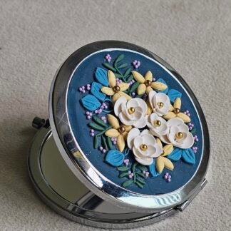 Handcrafted floral compact mirror with ivory roses, yellow petals, teal leaves, and lilac accents on a deep blue base in a silver-tone case