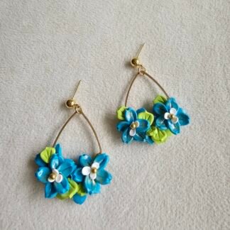 Handcrafted polymer clay blue floral earrings with green leaf accents and gold arc frame on a neutral fabric background
