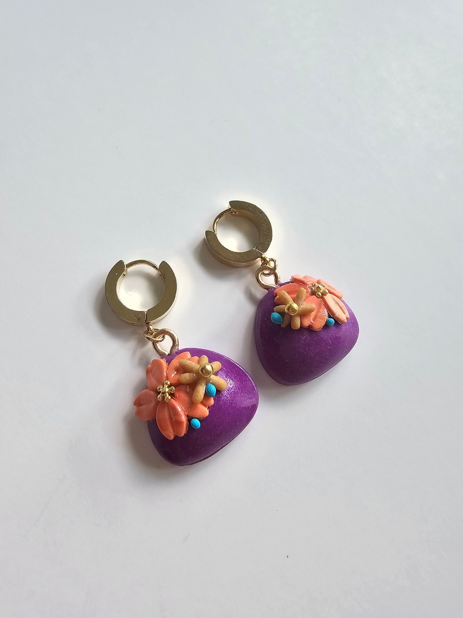 Gold plated huggie earrings with violet polymer clay puff charms and peach floral details on a white background