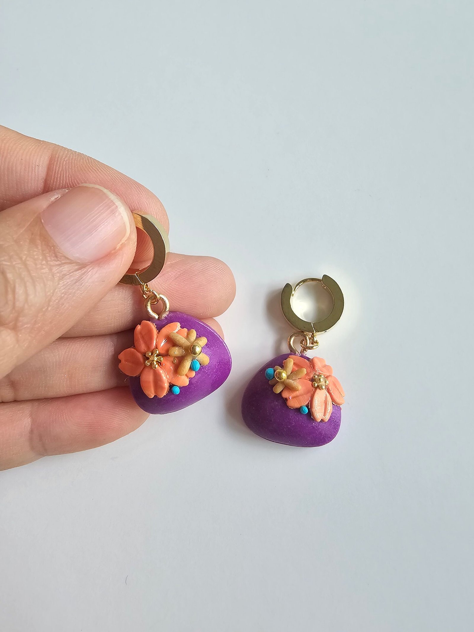 Gold plated huggie earrings with violet polymer clay puff charms and peach floral details on a white background