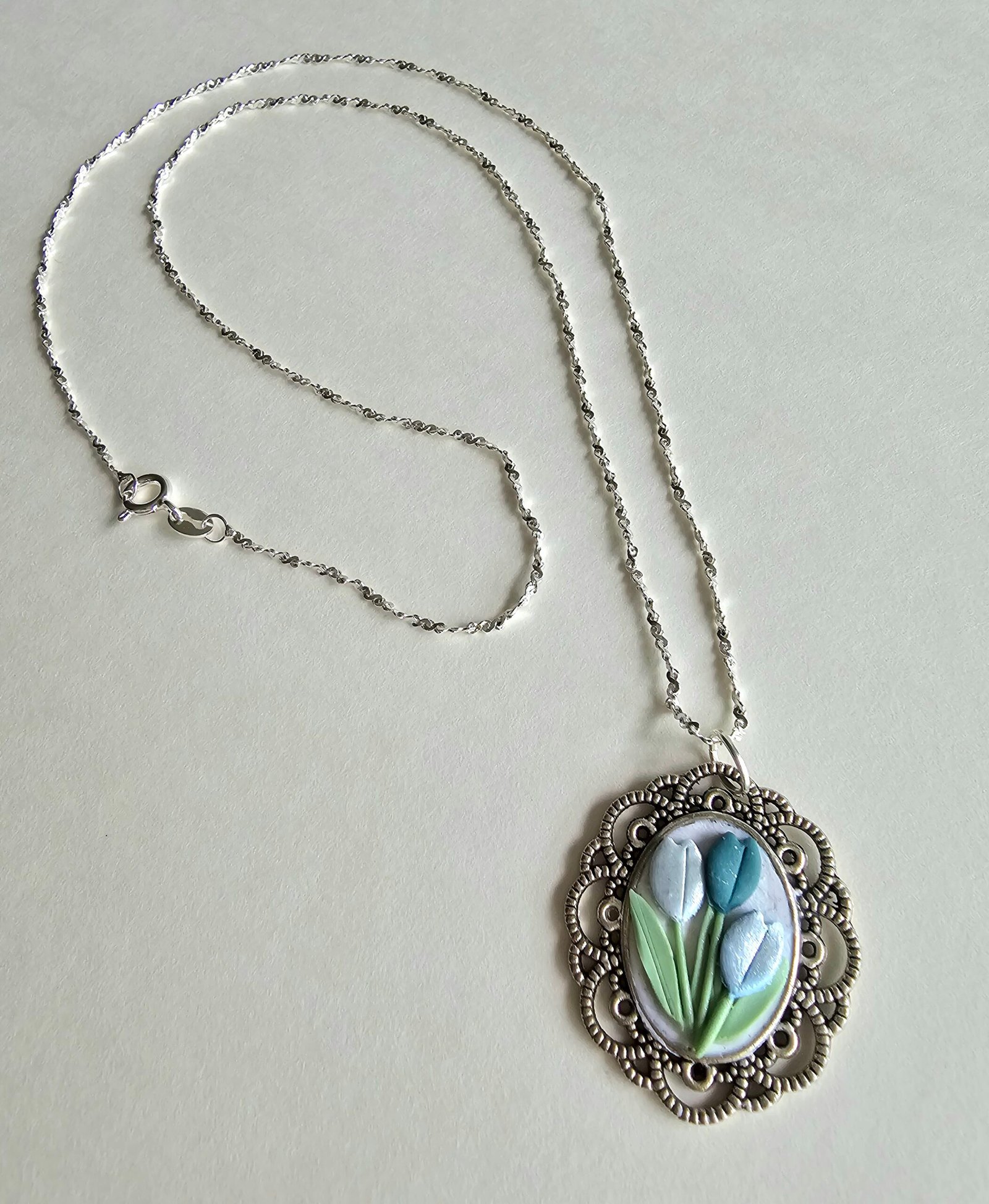 Handcrafted polymer clay floral pendant necklace in pastel blue and green, set in an ornate silver oval frame, on a 925 sterling silver chain by Haeng Bok Clay