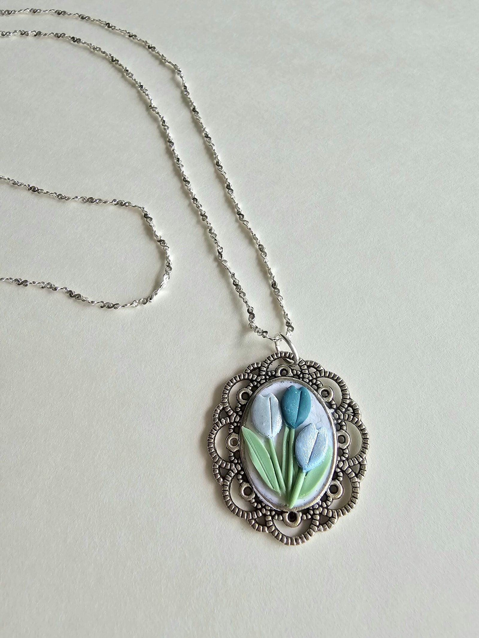 Handcrafted polymer clay floral pendant necklace in pastel blue and green, set in an ornate silver oval frame, on a 925 sterling silver chain by Haeng Bok Clay