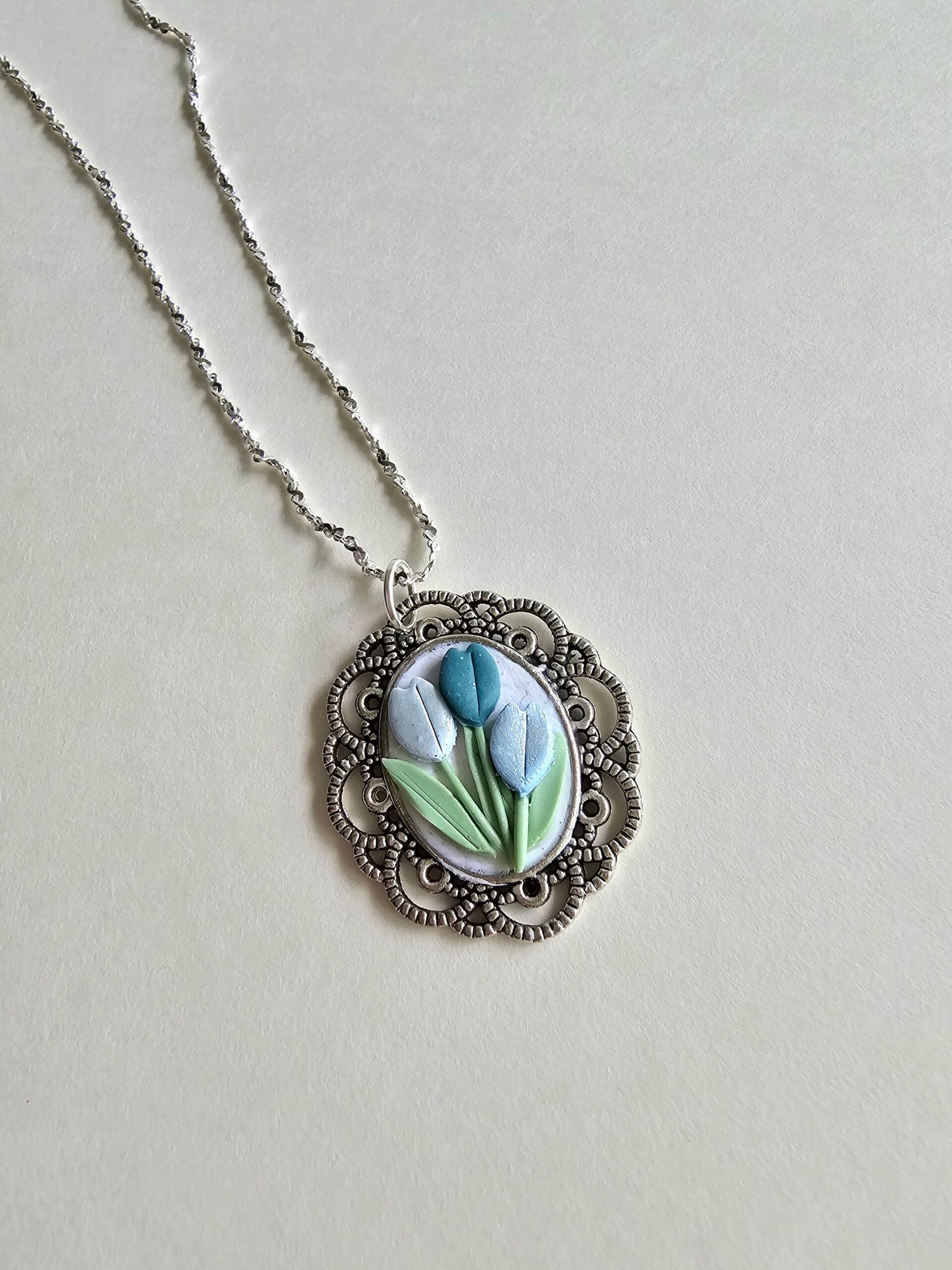 Handcrafted polymer clay floral pendant necklace in pastel blue and green, set in an ornate silver oval frame, on a 925 sterling silver chain by Haeng Bok Clay