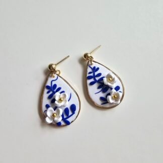Handcrafted polymer clay teardrop earrings with blue-and-white porcelain-style floral detailing, layered white clay flowers with gold centres, and polished gold ball stud posts, photographed on a clean neutral backdrop.