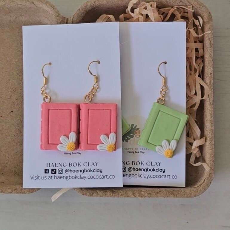 Handcrafted polymer clay mini book earrings in soft pink and sage green, each detailed with a small white daisy charm and finished with gold-toned hooks, displayed on Haeng Bok Clay branding cards in eco-friendly packaging.