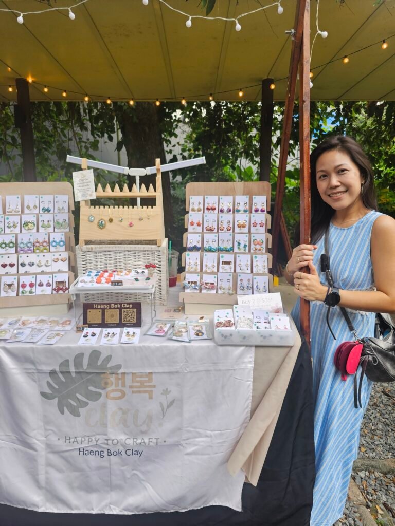 Founder of Haeng Bok Clay standing beside her handmade polymer clay jewellery booth at Sentosa Festive Market, Palawan Beach, featuring colourful floral earrings and accessories.